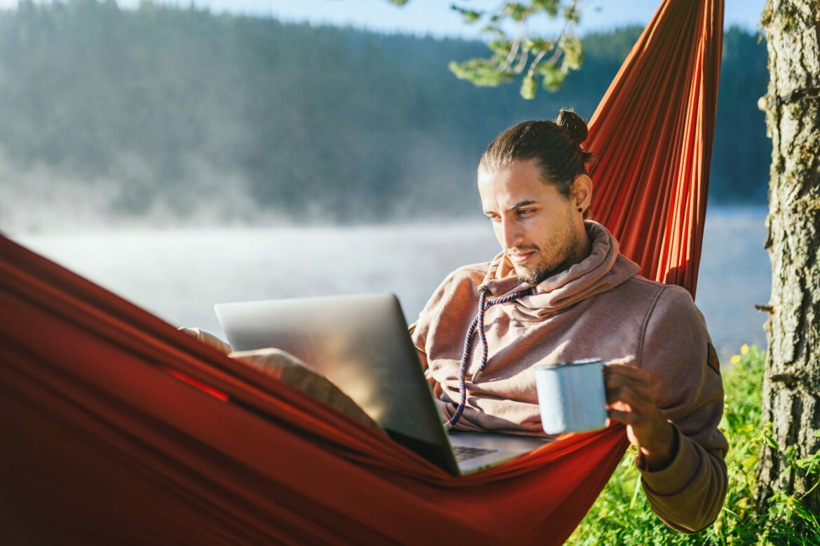 1966d5a066466c4678a58651e10fb33b Young man in hammock working on his laptop and drinking hot coffee. Leisure activities / Remote working concept.