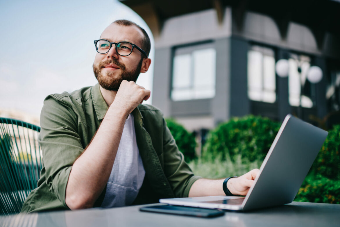 Young handsome male 20 years old in eyeglasses sitting at street cafe during free time with new modern laptop, bearded man freelancer  thoughtfully looking aside and thinking about distance work